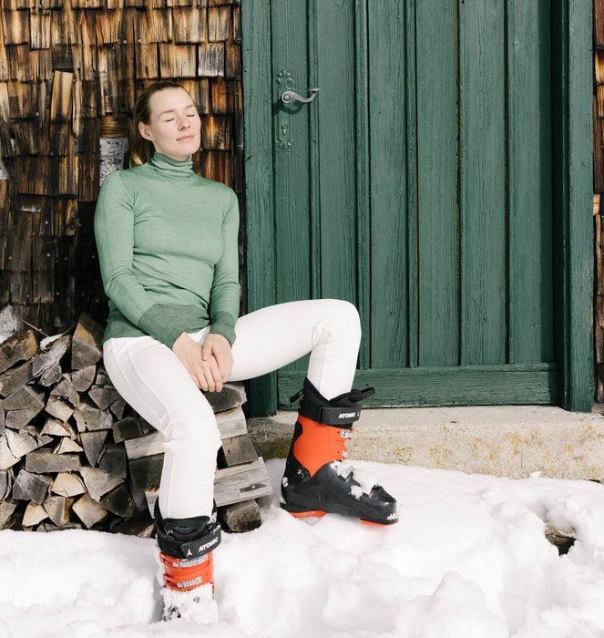 A lady in skiwear sits leaning against a wooden hut in the sun after skiing