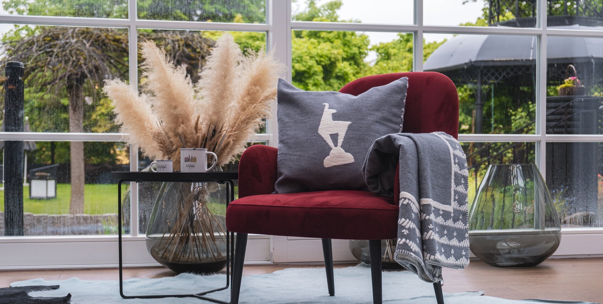 Ein Sessel mit einem grauen Kissen mit Gams Aufdruck und schönem grünen Garten im Hintergrund